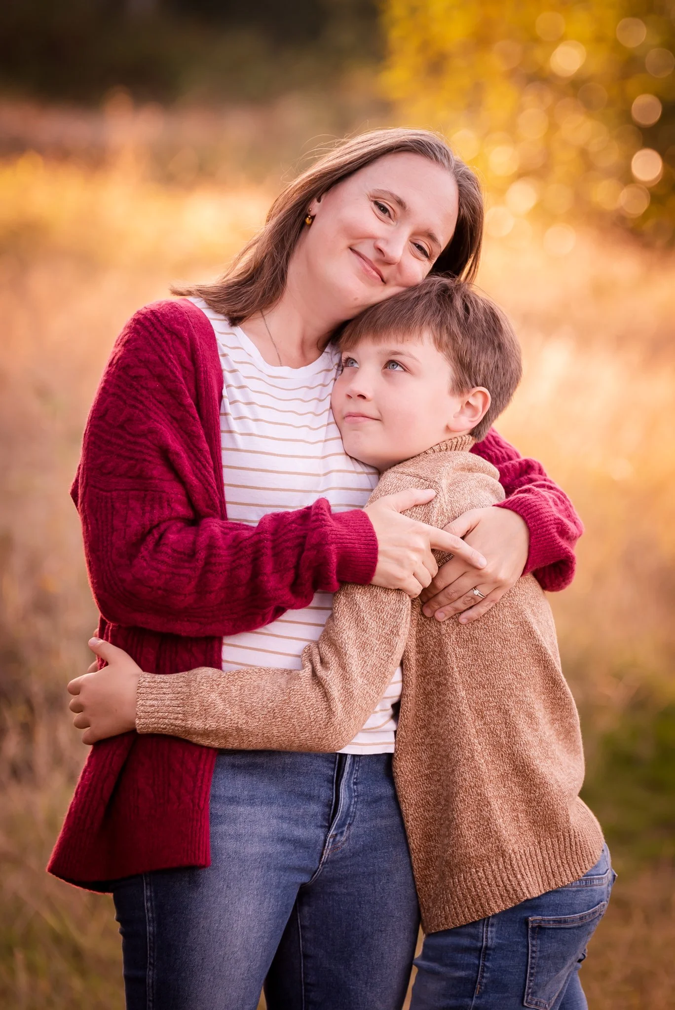 golden yellow grass and trees in background. mom in white and tan striped shirt with red cardigan sweater and jeans hugs school age son wearing tan sweater and jeans and hugging her back while looking up at her
