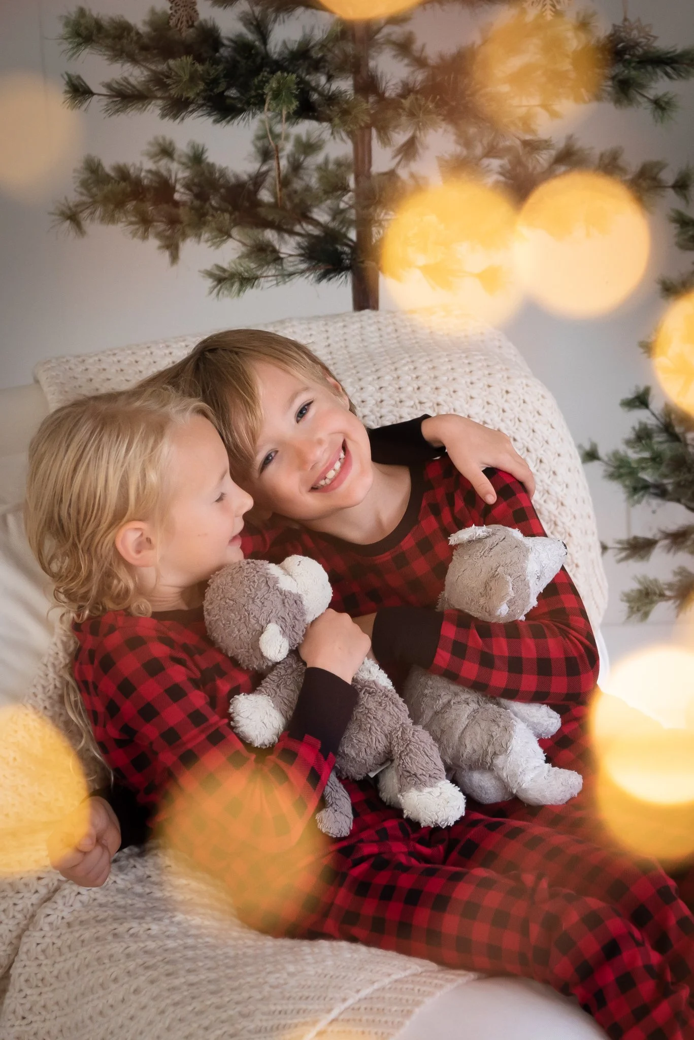 school age brother and sister snuggle on a cream chair with each other and their stuffies. Kids are in red and black buffalo check jammies with evergreen trees behind and twinkle lights in the foreground.