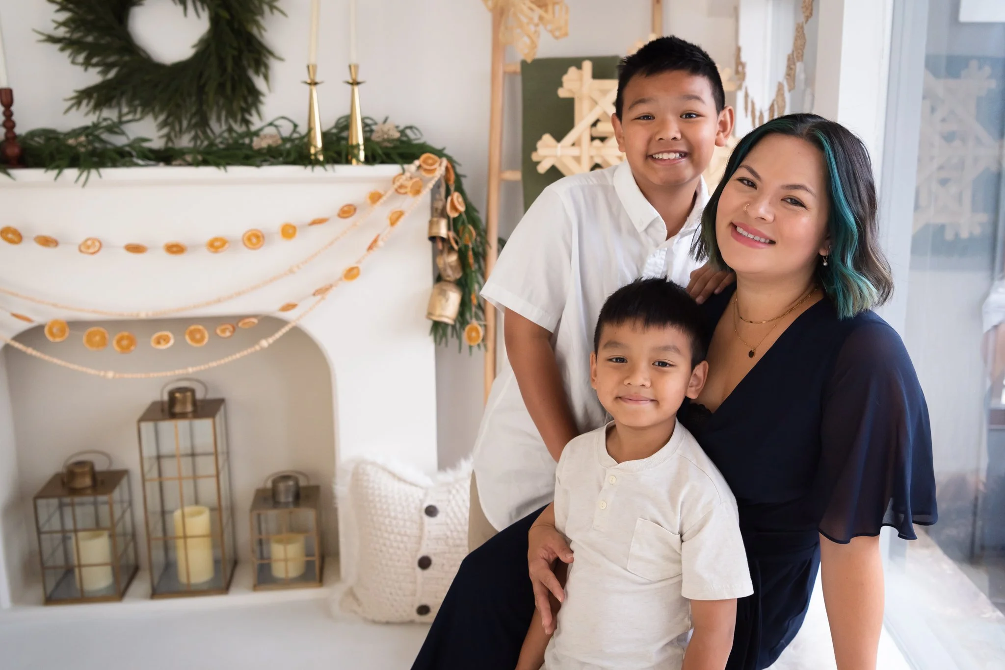 Mom and her two boys sit by the window with a holiday fireplace set behind them. Set is in greens, beige, and whites. Mom and boys are in white and navy dress clothes