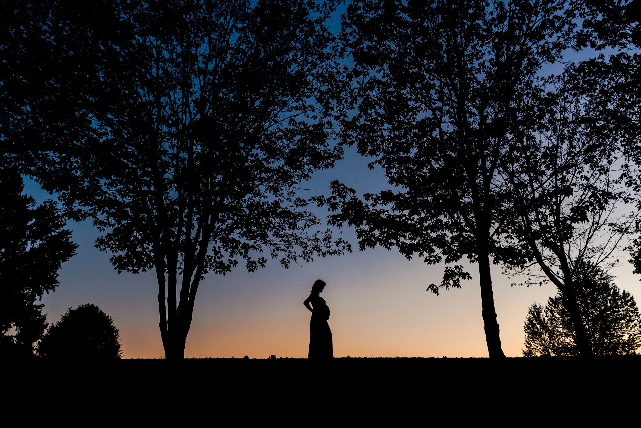 sunset silhouette of pregnant woman surrounded by trees. Sky is orange down around her transitioning to dark blue a the top of the image.
