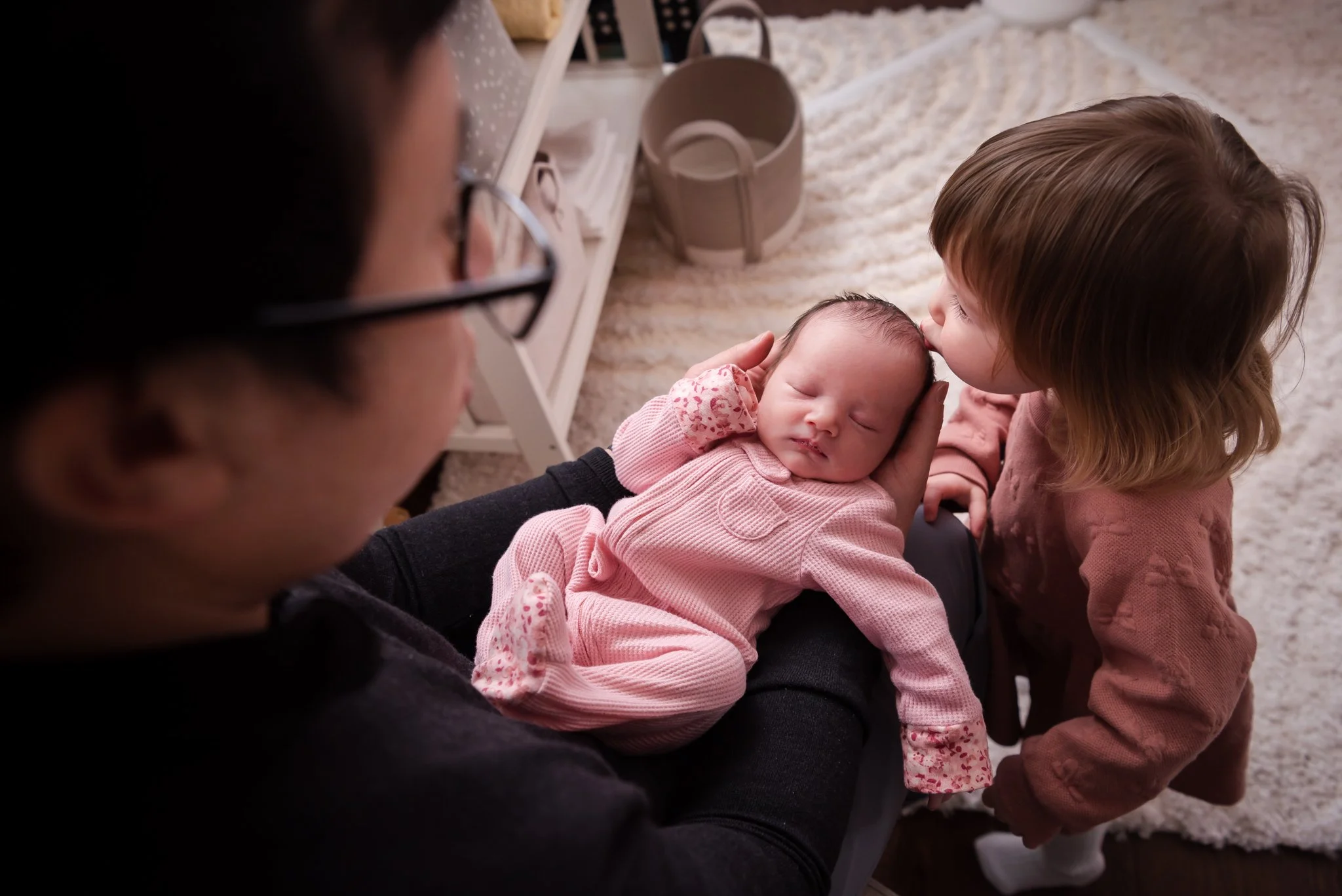 looking down at baby girl in dad's arms while toddler sister kisses her new baby sister on the head. Girls in pink with cream carpet behind them.