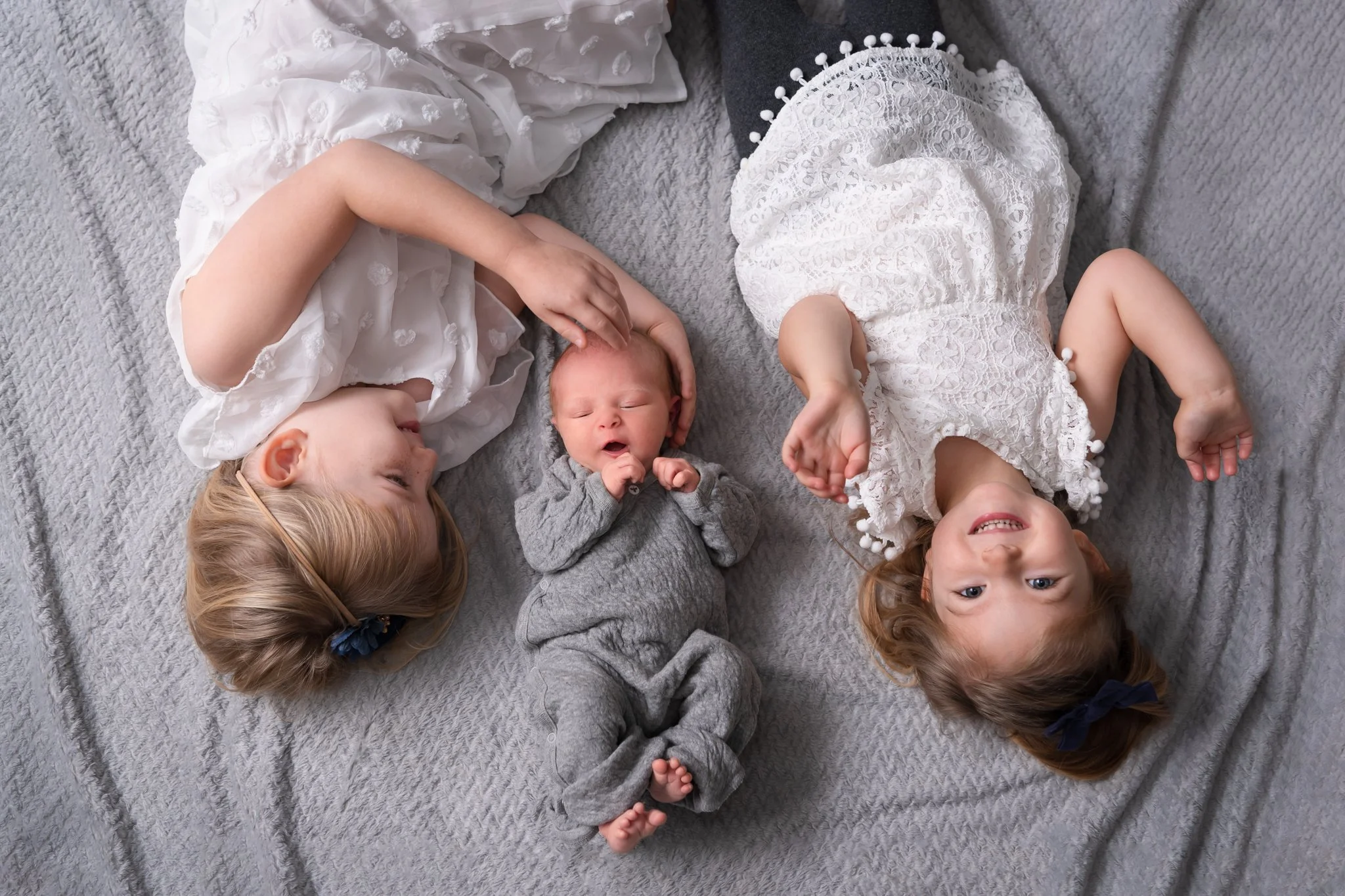 two older sisters laying on a gray blanket with baby brother in between them. Oldest sister is gently cradling brother's head. Middle sister is smiling up at the camera. Sisters in white dresses