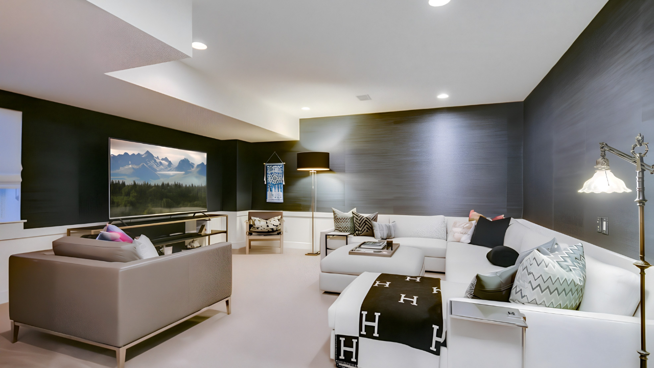 Modern living room with white sectional sofa, armchair, and various decorative pillows. A large flat-screen TV on the wall, floor lamp, and another standing lamp provide lighting. A window with a landscape view and a wall hanging are also visible.