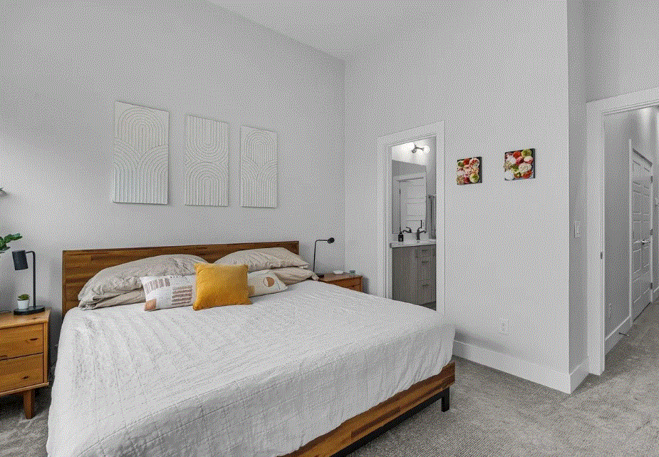 A neatly made bed with beige and mustard pillows in a minimalist bedroom with white walls, a wood nightstand with a small plant and lamp, and a small attached bathroom visible through a door.