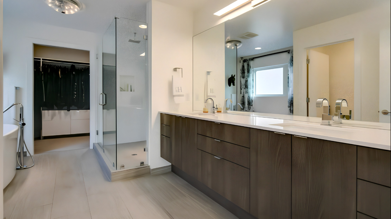 Modern bathroom with a long white countertop, dark wood cabinets, large mirror, double sinks, in a brightly lit space with a window and shower enclosure.