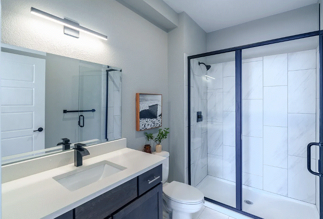 Modern bathroom with a white vanity, black faucet, toilet, a framed artwork, a small potted plant, and a glass-enclosed shower with white tiles and black hardware.