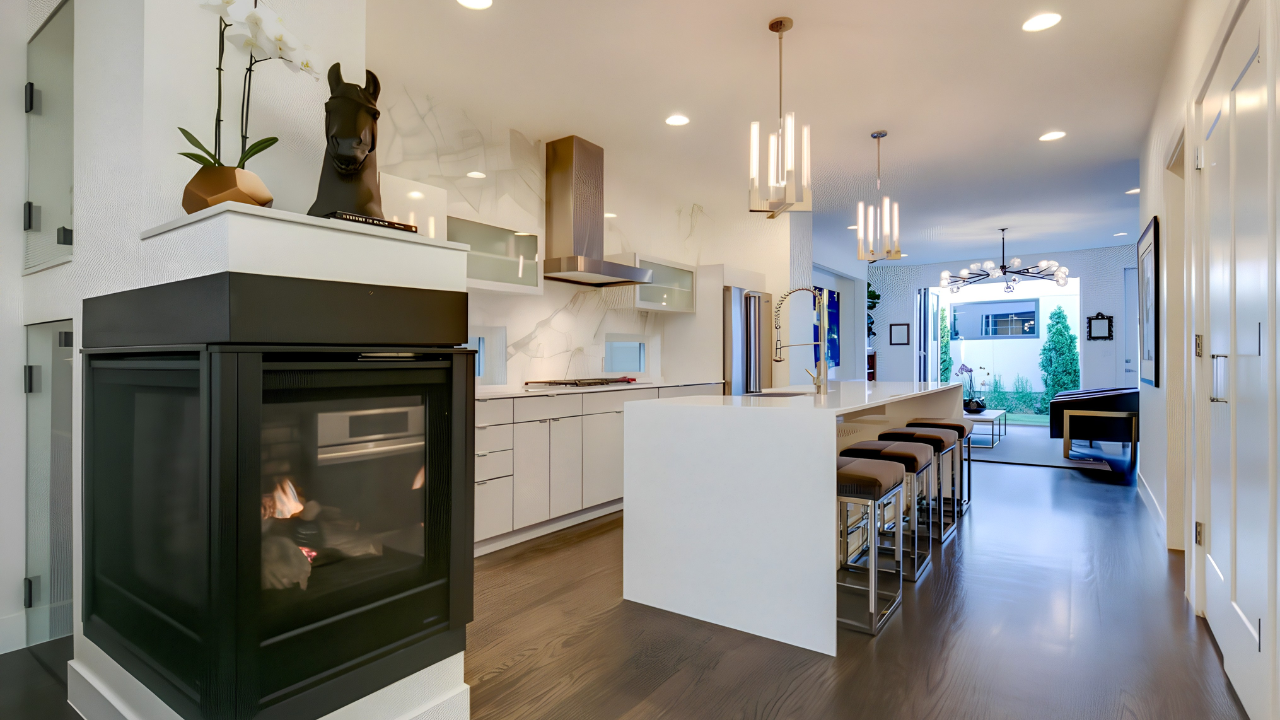 Modern open-concept kitchen and living room with white cabinetry, a kitchen island with bar stools, contemporary lighting, and a view of the backyard garden.
