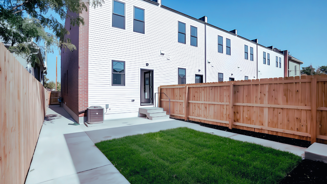 Backyard scene with a small patch of green grass, concrete sidewalk, wooden fence, and white modern townhouse with multiple windows and steps leading to a door.