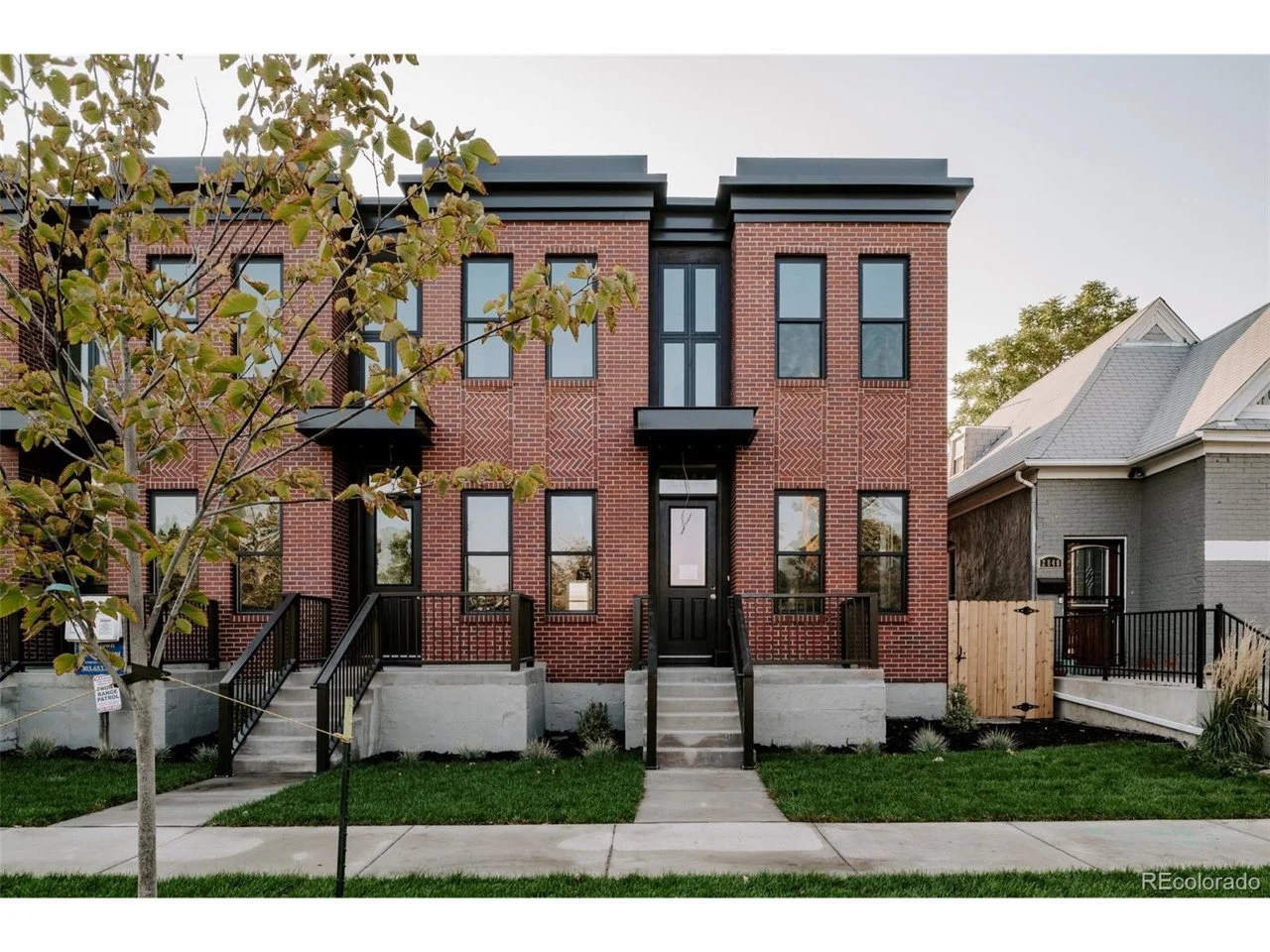 A modern three-story brick townhouse with black framed windows, a small front step, and a path leading to the sidewalk.