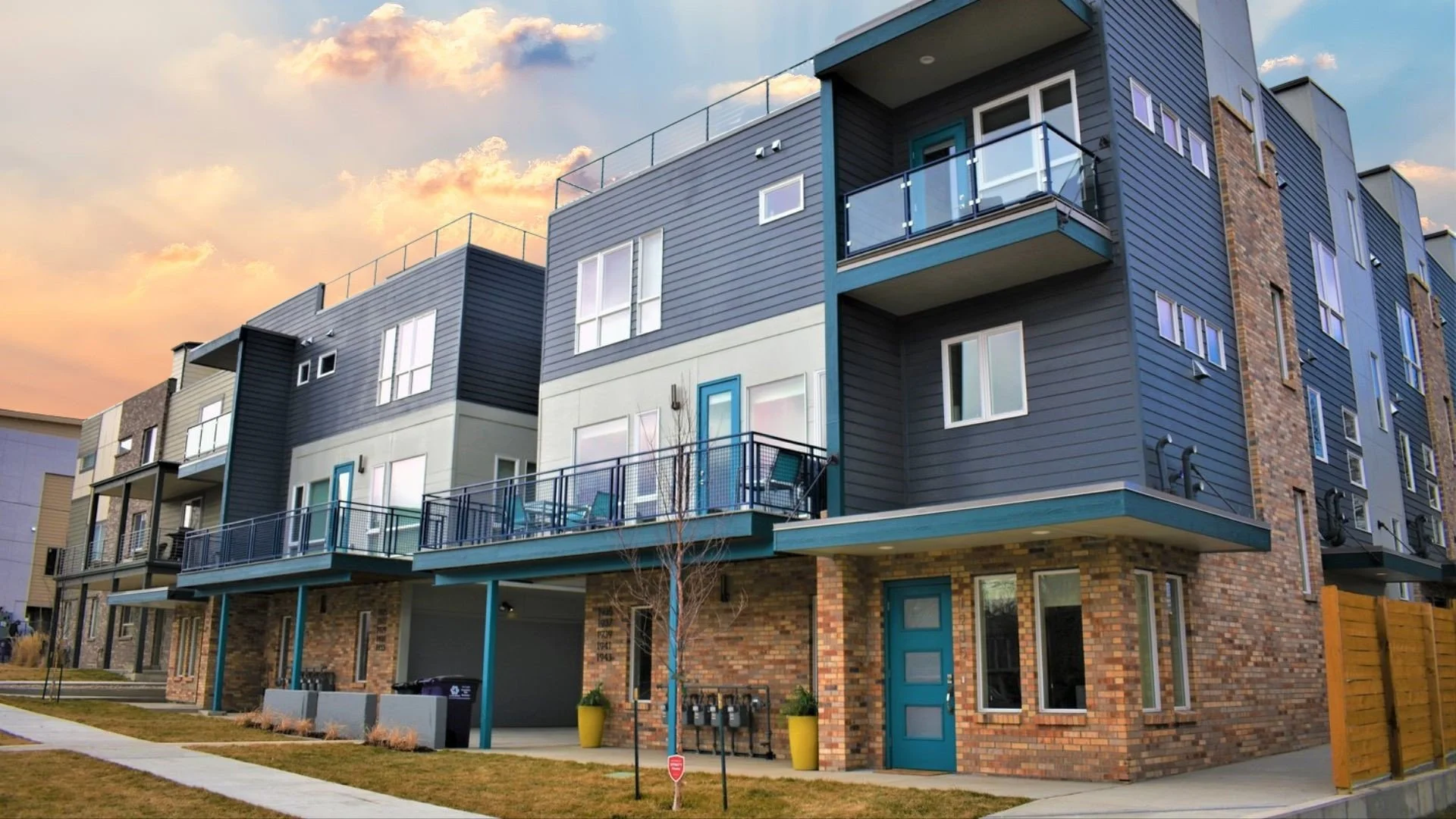 Modern multi-story apartment building with brick and siding exterior, balconies, large windows, and a blue door, at sunset.