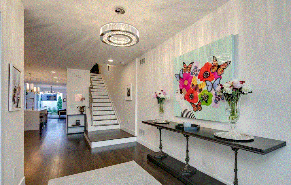 Interior of a modern house with stairs and a hallway. There is a large colorful butterfly and floral artwork on the wall above a dark console table with vases of flowers. The space has hardwood floors and light-colored walls.