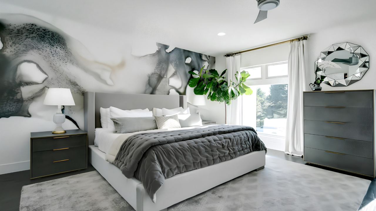 Modern bedroom with gray and white decor, large abstract wall art, gray bed with multiple pillows, gray bedside table with lamp, gray dresser with decorative mirror, window with white curtains, green plant, and light-colored carpet.