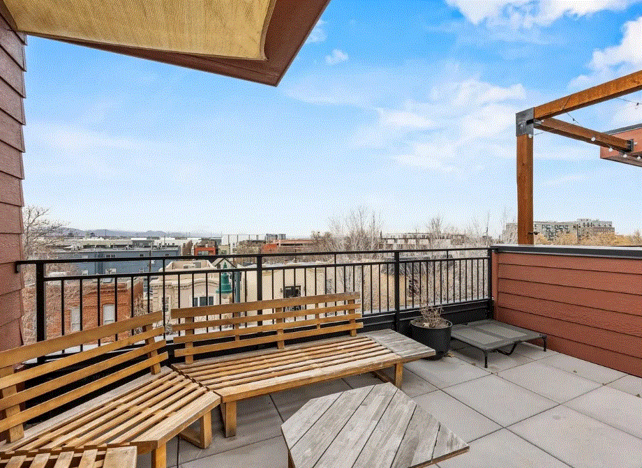 Rooftop balcony with wooden seating, potted plant, and view of concrete buildings under a blue sky.