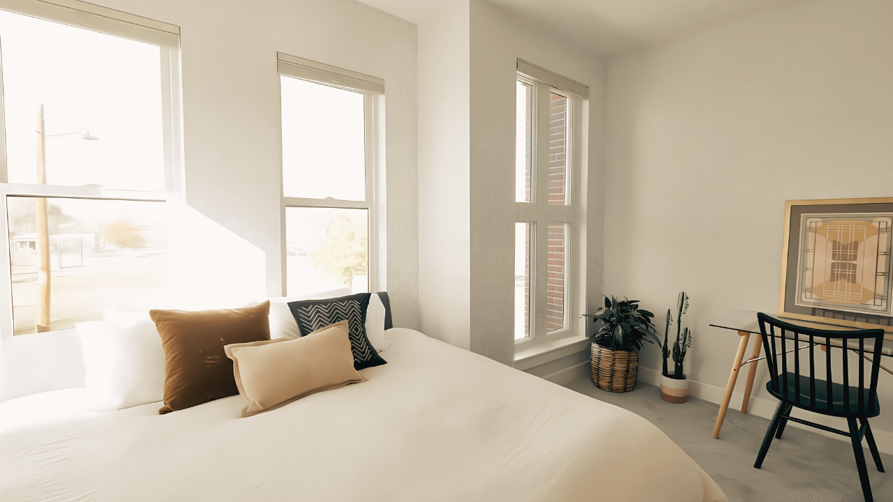 A bright, modern bedroom with large windows, a white bed with several decorative pillows, and a small desk with a black chair. There are two potted plants in the corner and a framed artwork on the wall.