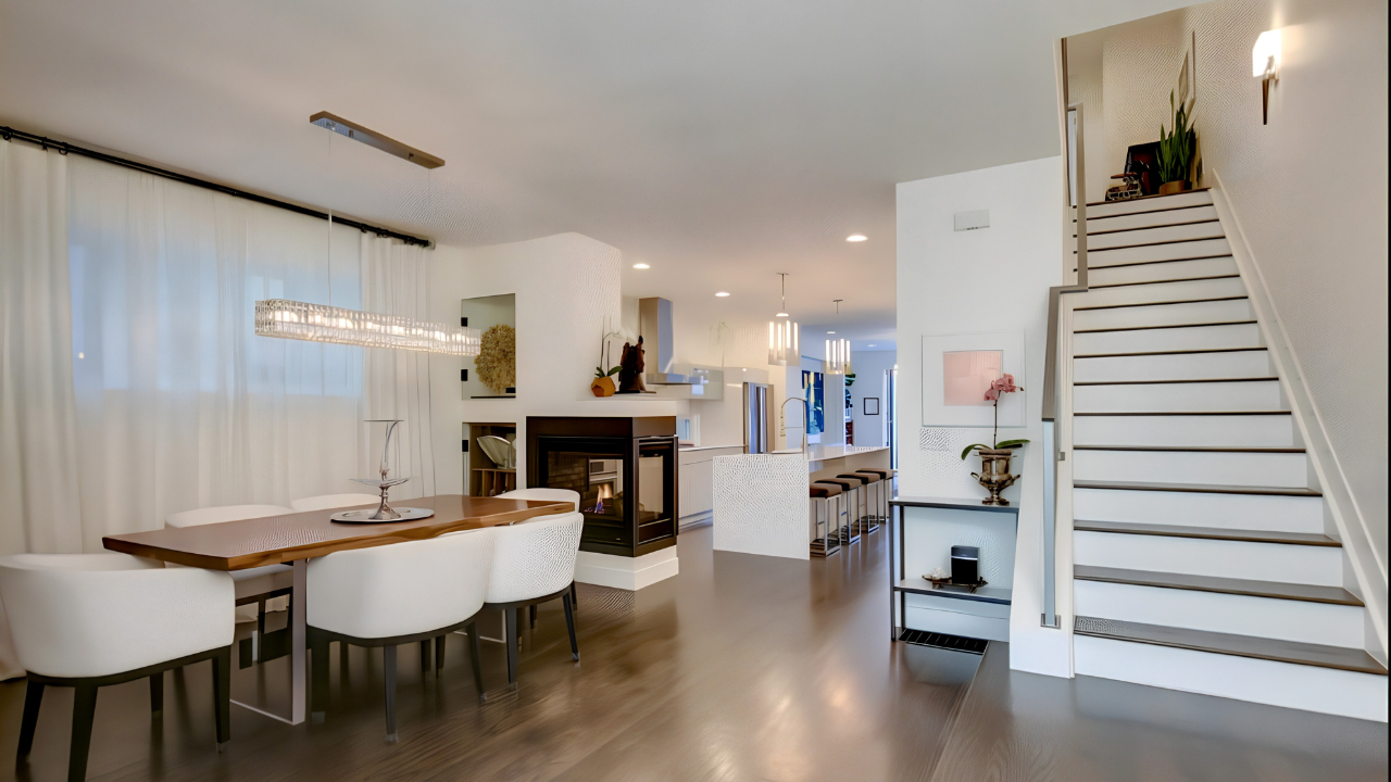Modern open-concept living and dining area with a staircase on the right, a dining table with seven white chairs, a fireplace in the middle, and a kitchen with a breakfast bar in the background.