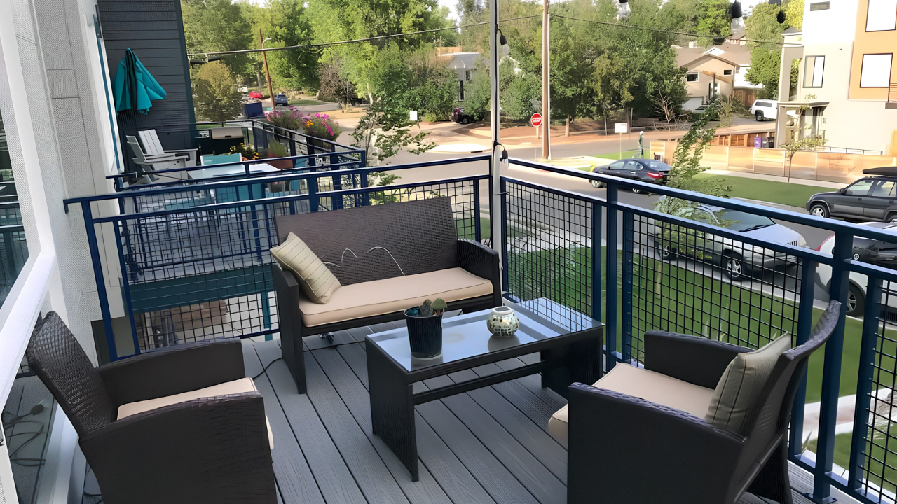 View of a balcony with outdoor seating, including a black wicker sofa and two matching armchairs with beige cushions, a black coffee table with decorative items, and a view of a residential street with cars, trees, and apartment buildings.