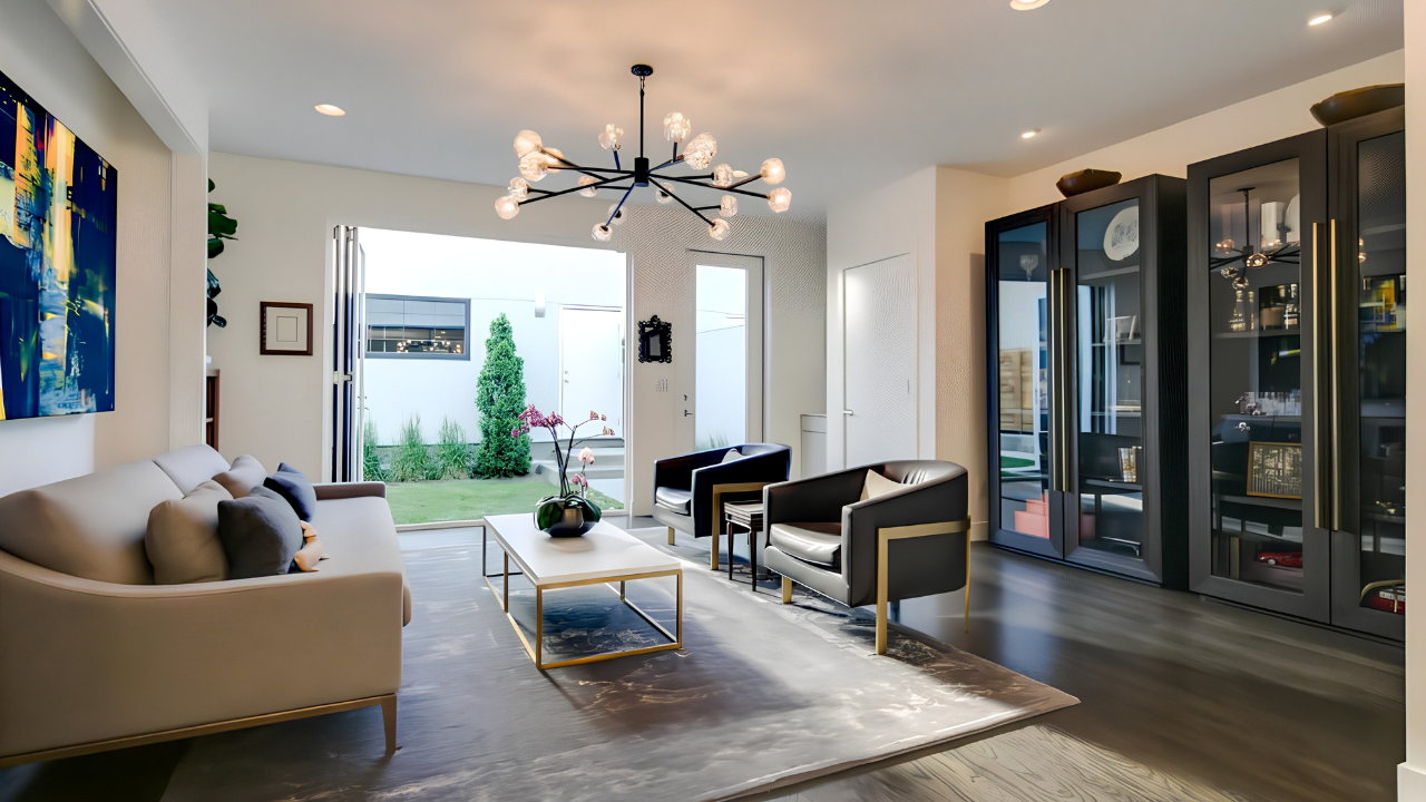Modern living room with a white sofa, black and white armchairs, glass front cabinets, modern chandelier, and sliding glass doors leading to a backyard garden.