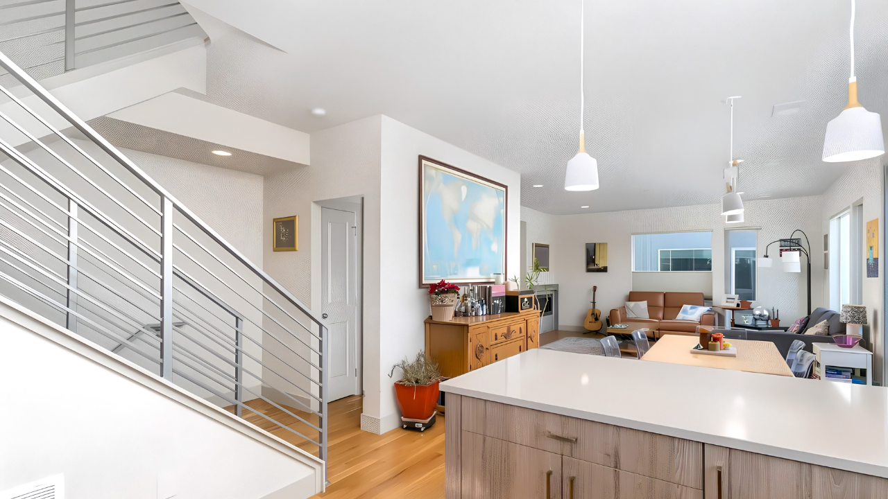 Open-concept living space with a modern staircase, white kitchen counter, and a brightly lit living room with a brown sofa, an armchair, a dining table, and musical instruments.