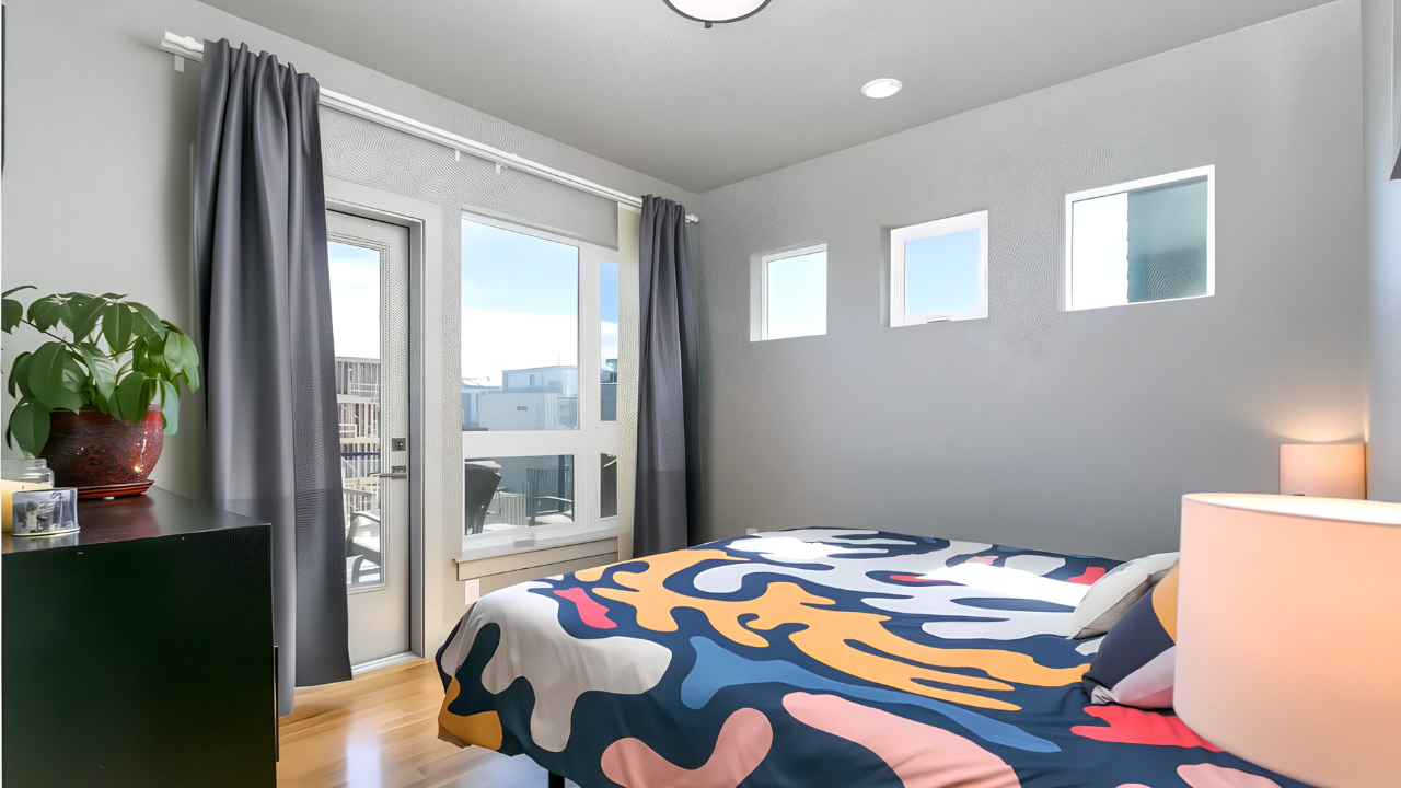 Bright bedroom with a bed that has a colorful patterned duvet, a nightstand with a lamp, a black dresser with a plant, and a large window with gray curtains and small windows on the wall, letting in natural light.
