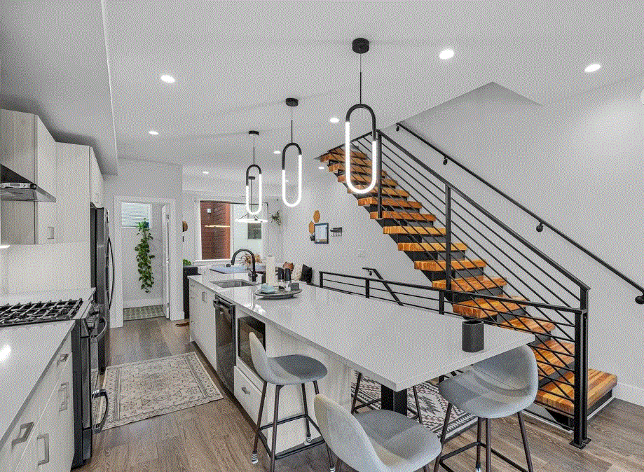 Modern kitchen with white cabinetry, a large island with seating, pendant lighting, and a staircase with wooden steps and black railing.