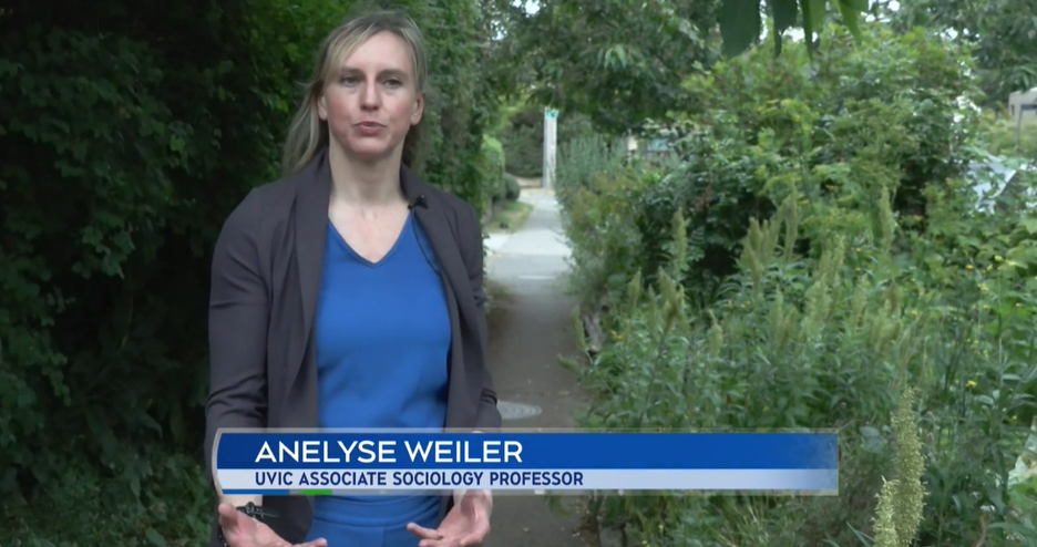 Anelyse Weiler, UVic Associate Sociology Professor, wearing a blue pantsuit and grey blazer and speaking with green foliage in background.