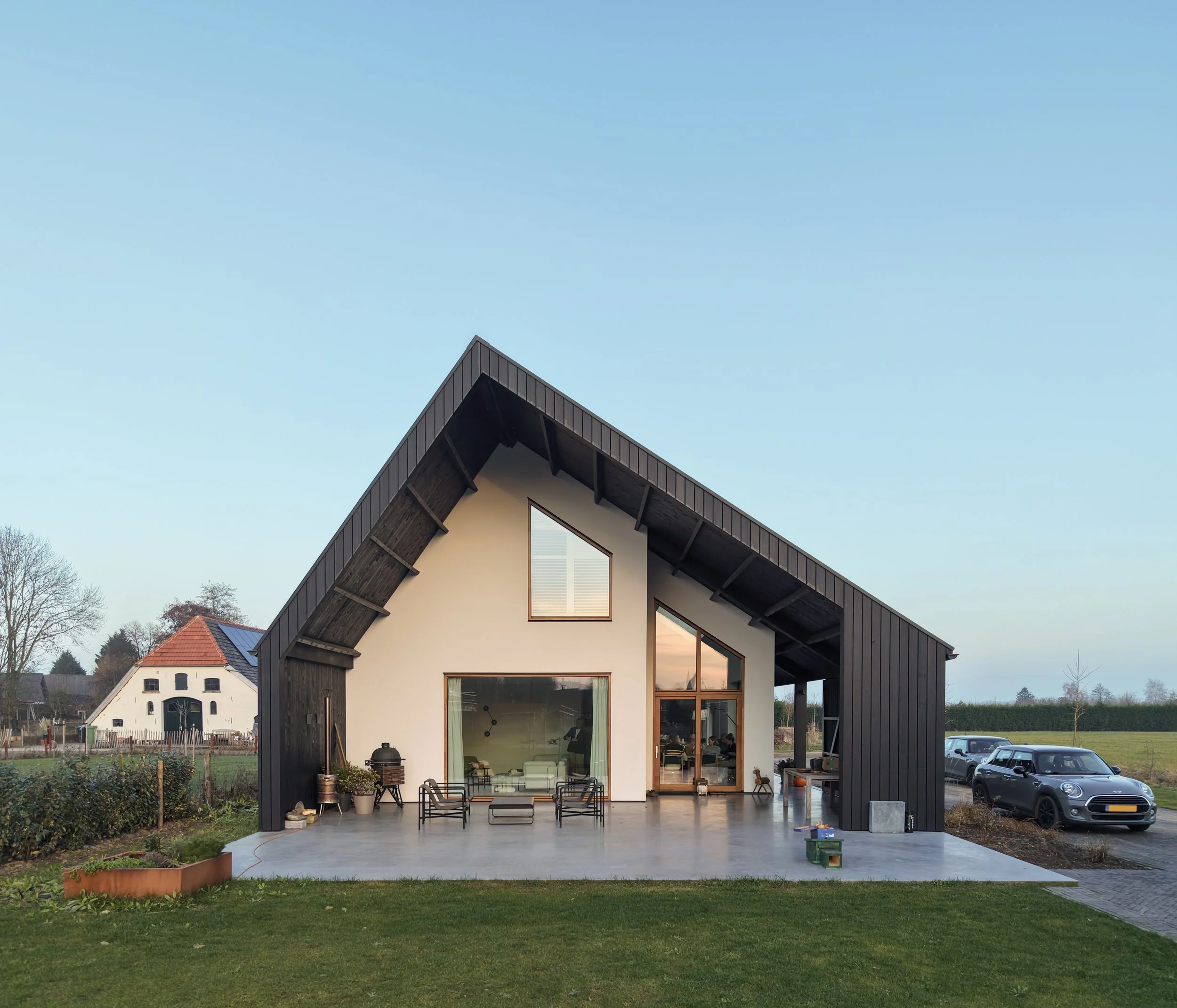 Achtergevel schuurwoning met wit gestucte gevel en houten kozijnen. Zwarte houten geveldelen met grote overkapping. Landelijke setting op voormalig boerenerf.