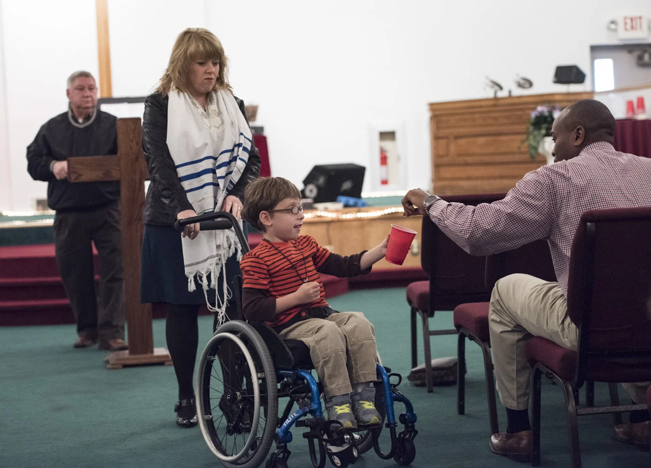  Josiah is taken around by his grandma Trish Anderson (left) to collect donations from church patrons in Dundar, W.Va. Steve Buckingham (right) puts money in Josiah’s cup. 