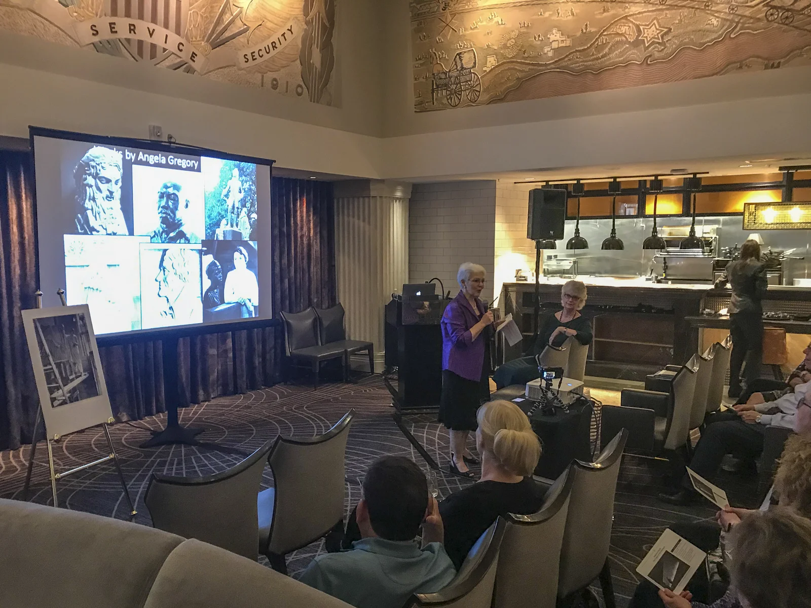 Susan Hymel, Angela Gregory historian, speaks in The Gregory restaurant, Watermark Hotel, Baton Rouge, at the launch of A Dream and a Chisel , 7 February 2019. Sculptured murals by Angela Gregory may be seen above.