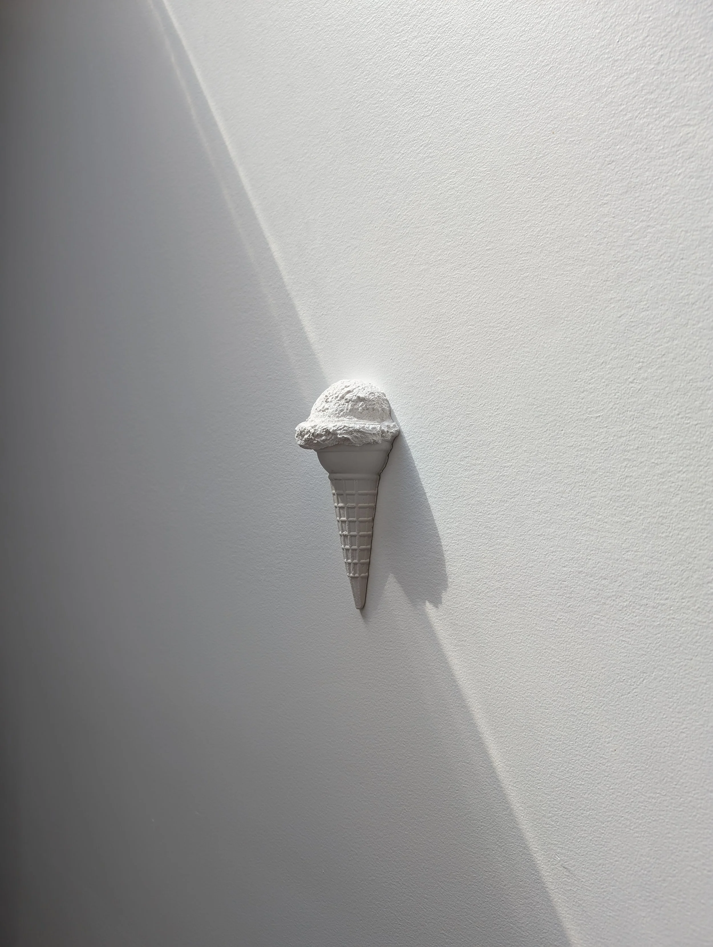 Minimalist white wall sculpture of a single ice cream cone, with contrasting light and shadows.