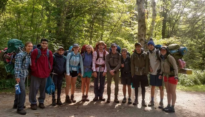 Group of hikers standing on a trail in a forest, with trees and sunlight in the background.