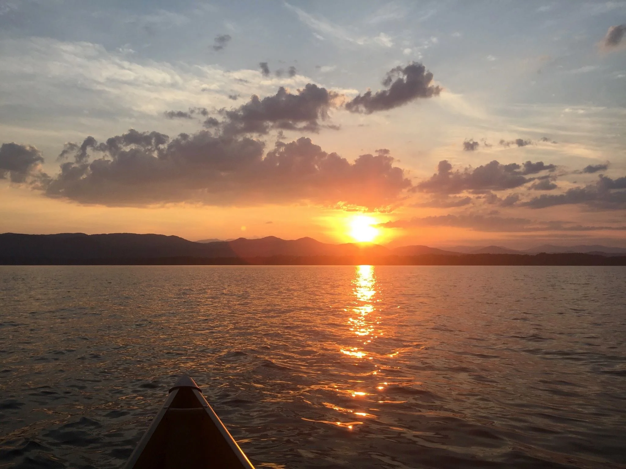 Sunset over a lake with dark clouds in the sky, mountains in the distance, and the tip of a canoe in the foreground.