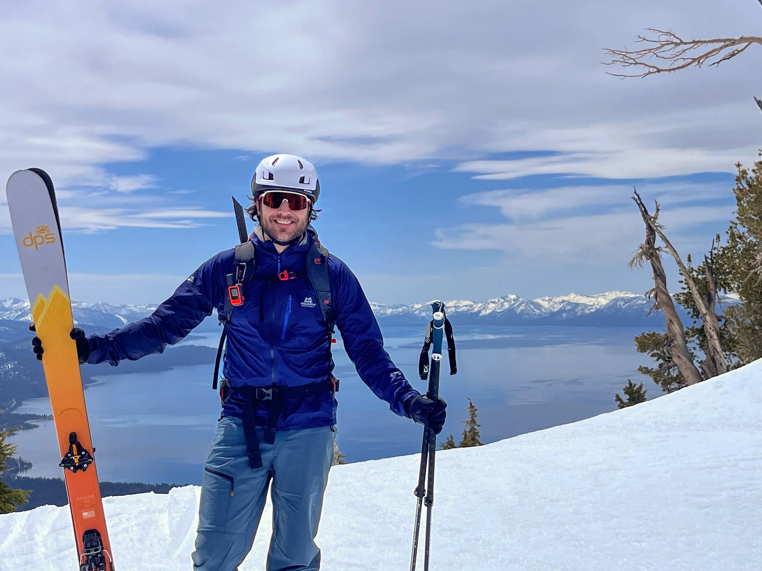 A man dressed in blue ski gear stands on snowy mountain, holding skis and poles, with snow-capped mountains and lake in the background.