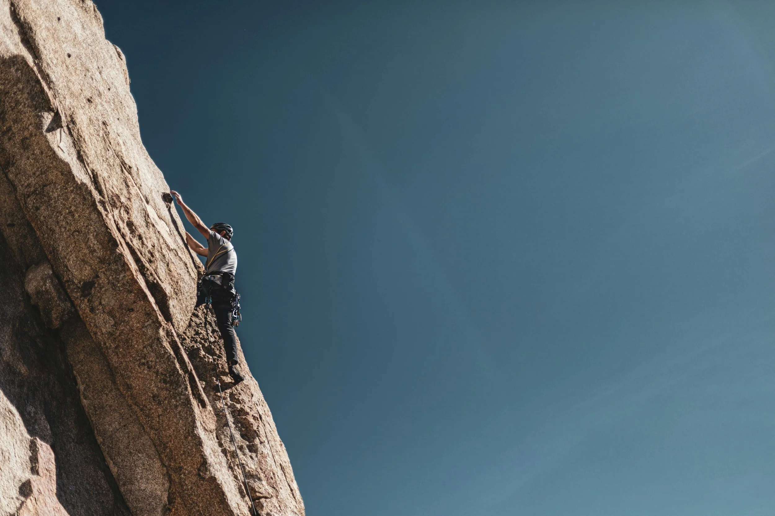 A person wearing a helmet and climbing gear is rock climbing a steep granite cliff against a clear blue sky.