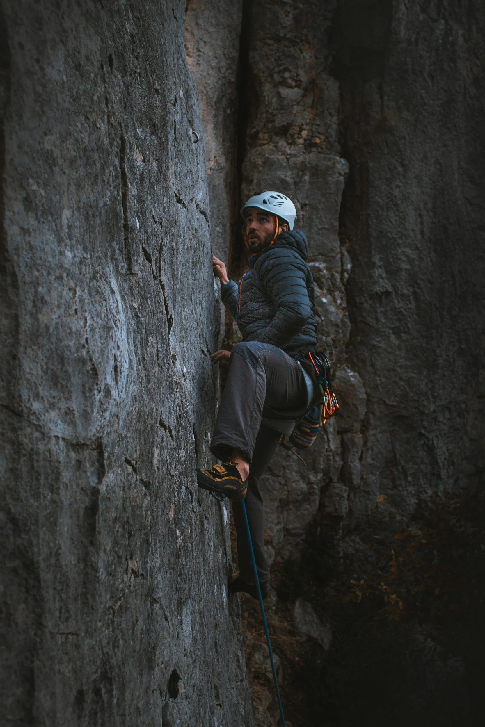 A man rock climbing on a vertical outdoor cliff, wearing a white helmet, black jacket, and climbing gear.