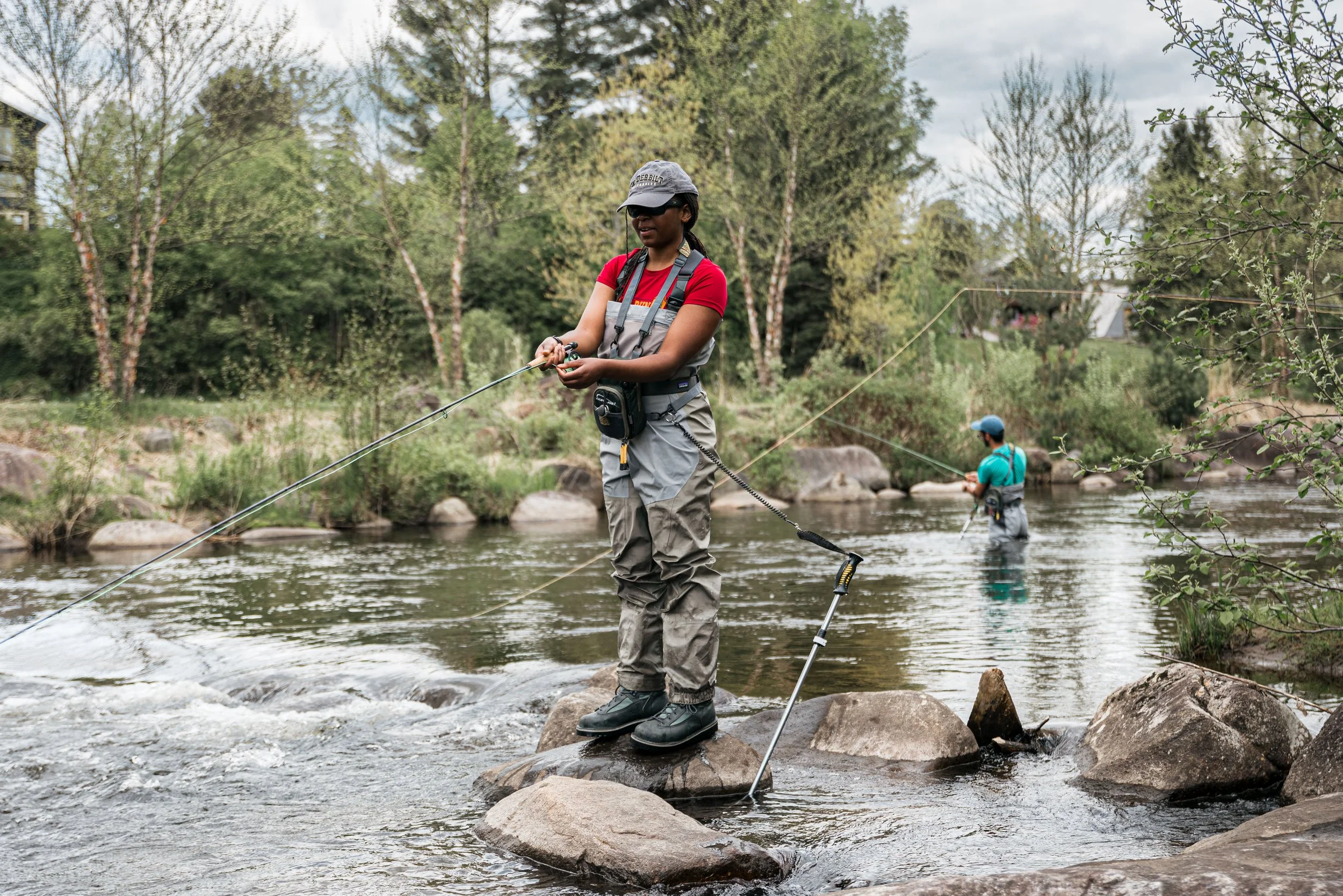 Two people fly fishing in a river surrounded by trees and rocks, wearing outdoor gear and hats.