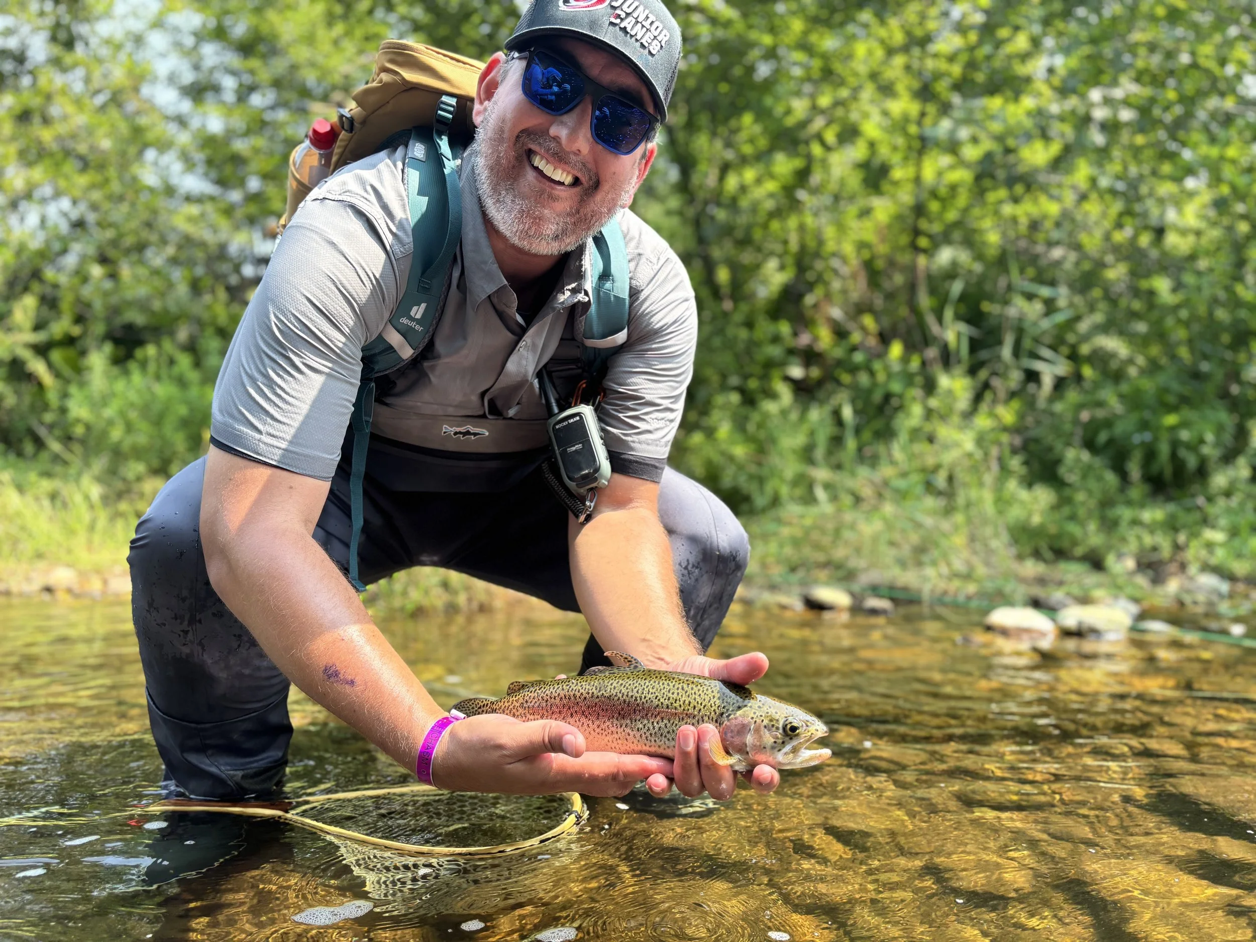A man wearing a gray cap, sunglasses, and a gray shirt crouches in a shallow stream while holding a colorful rainbow trout. He is smiling and has a backpack on his back.