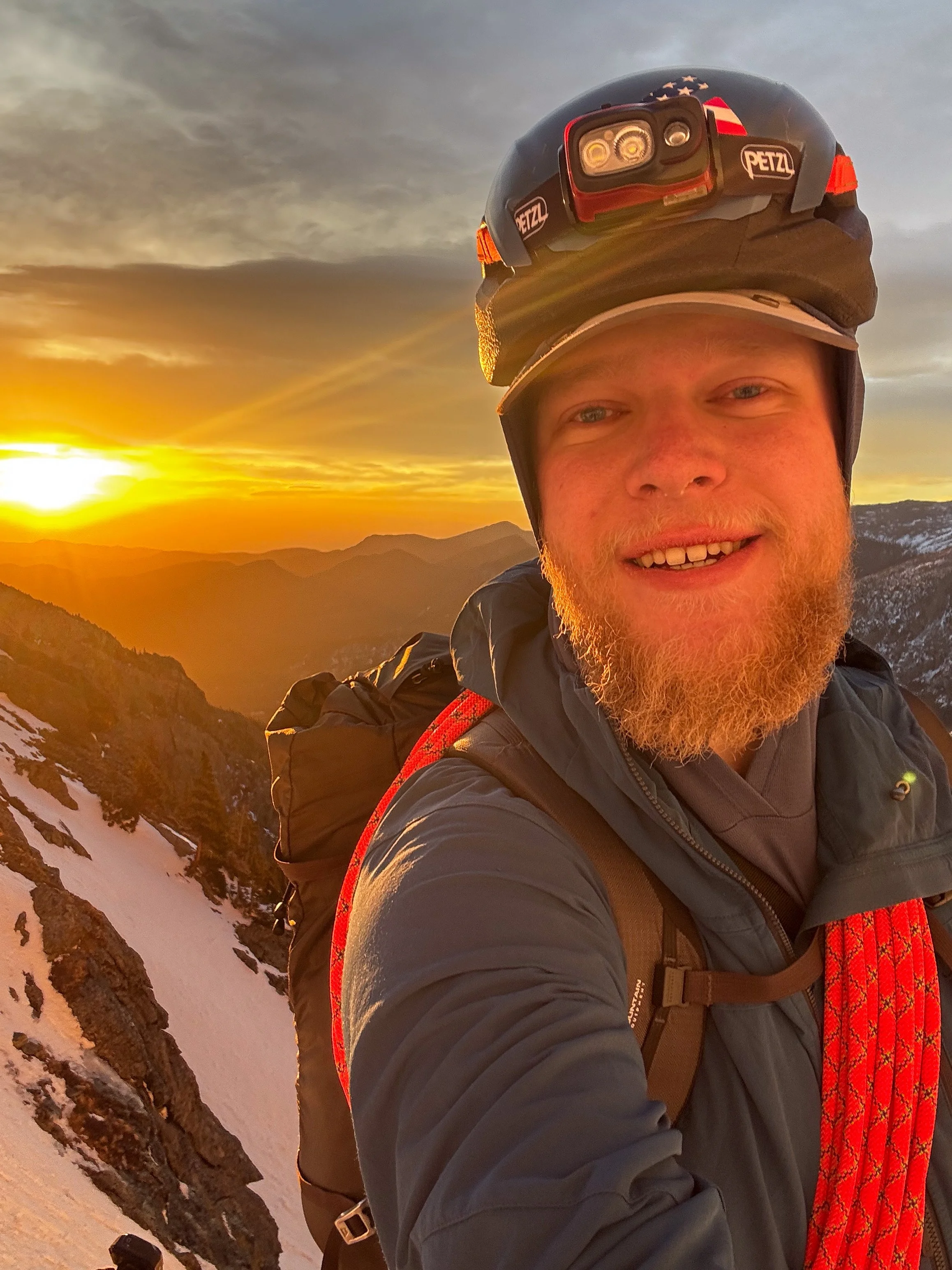 A man with a red beard and blue eyes taking a selfie on a snowy mountain during a sunset, wearing outdoor gear and a headlamp.