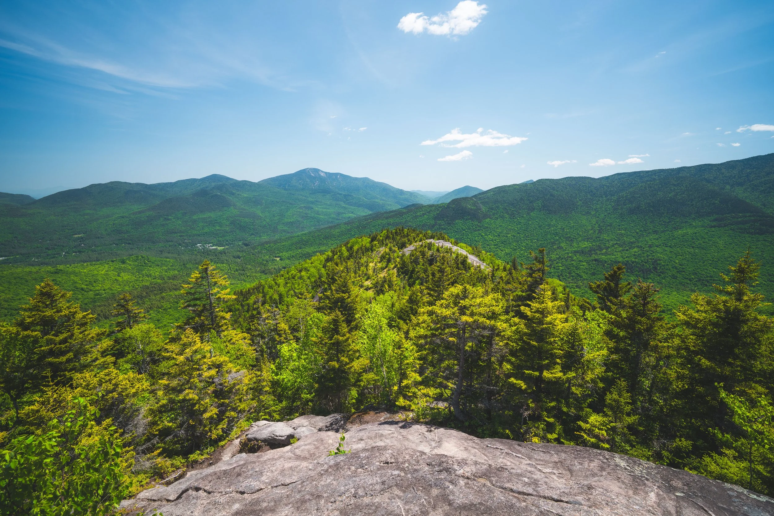 View from a rocky outcrop overlooking a lush green forest and mountain range under a bright blue sky with a few clouds.