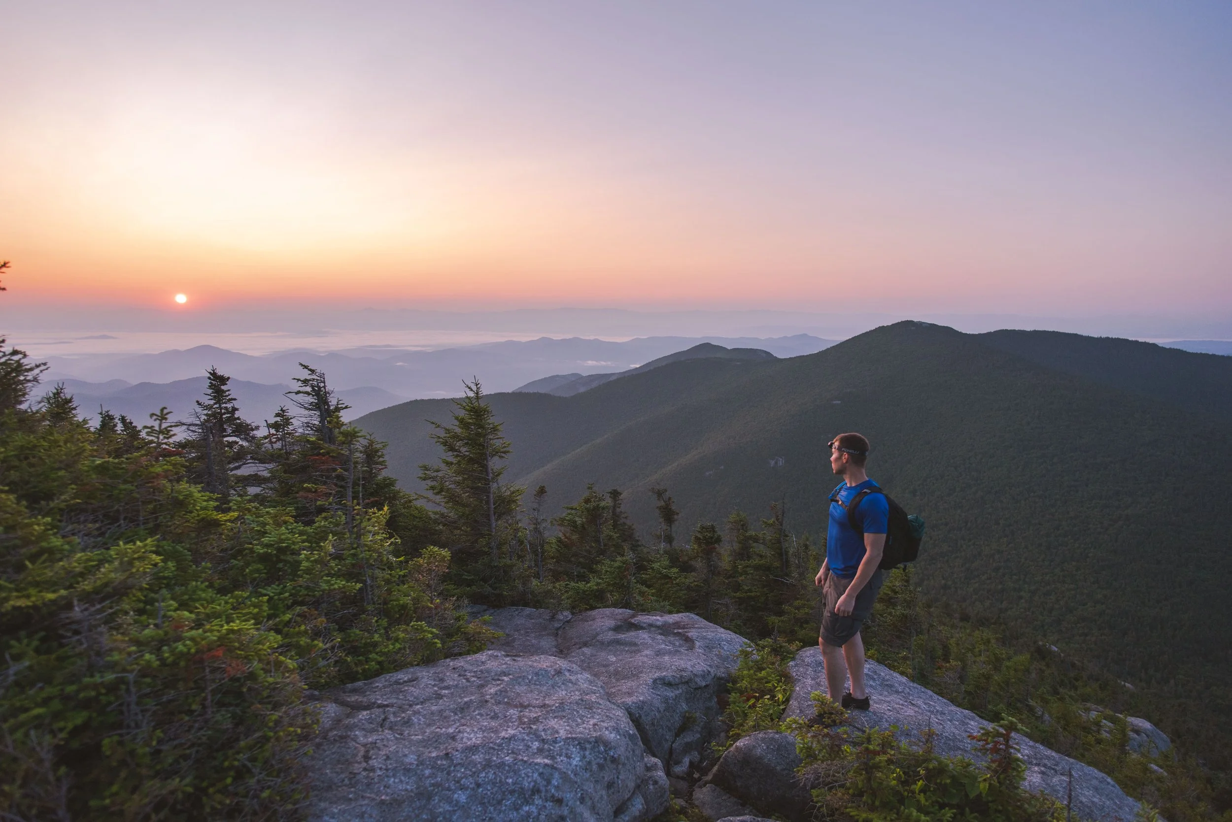 A man standing on rocks overlooking a mountain range during sunrise, wearing a blue shirt, shorts, and carrying a backpack.