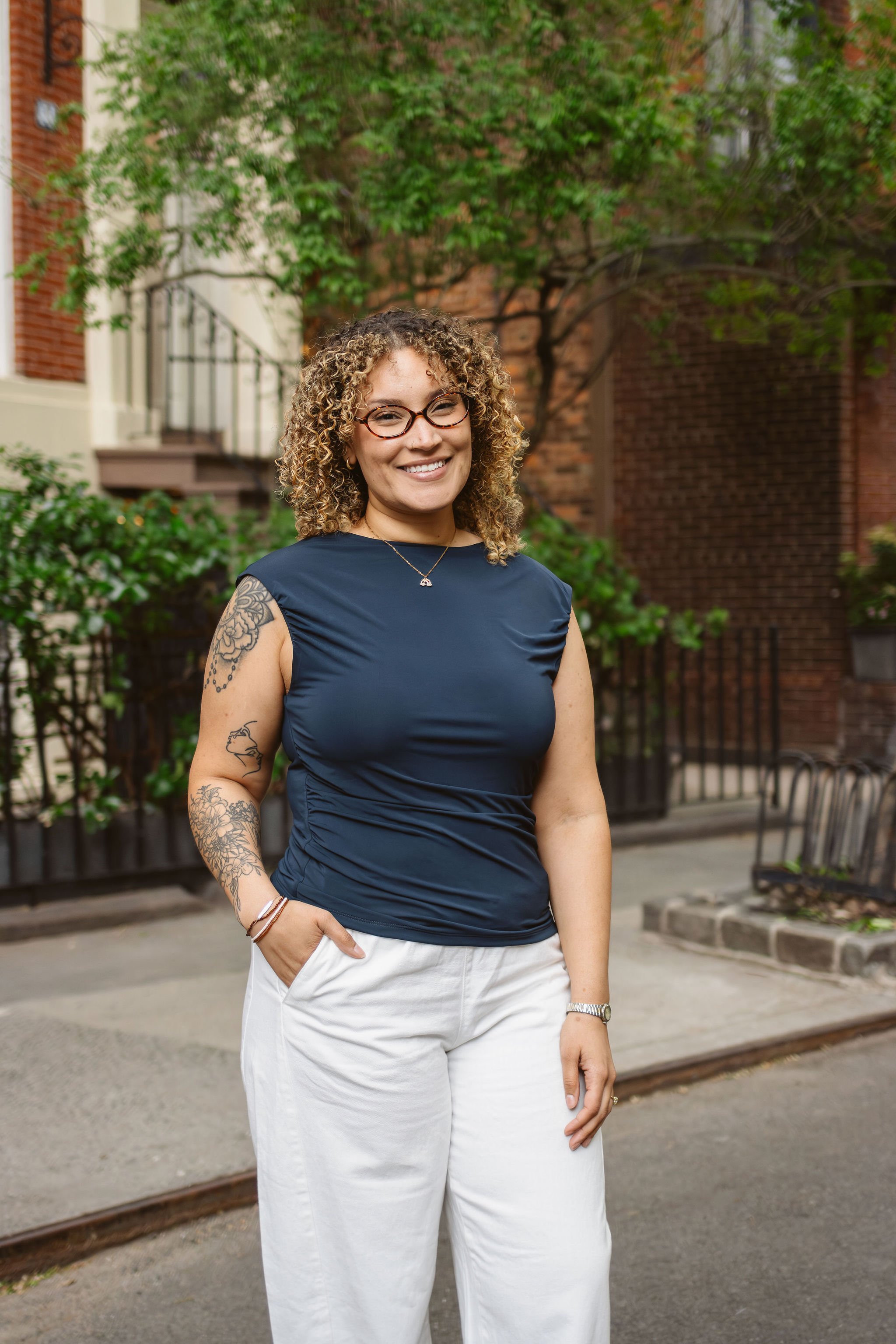 A woman with curly hair smiling outdoors in front of a bush, with a pink building and windows with window boxes filled with flowers in the background.