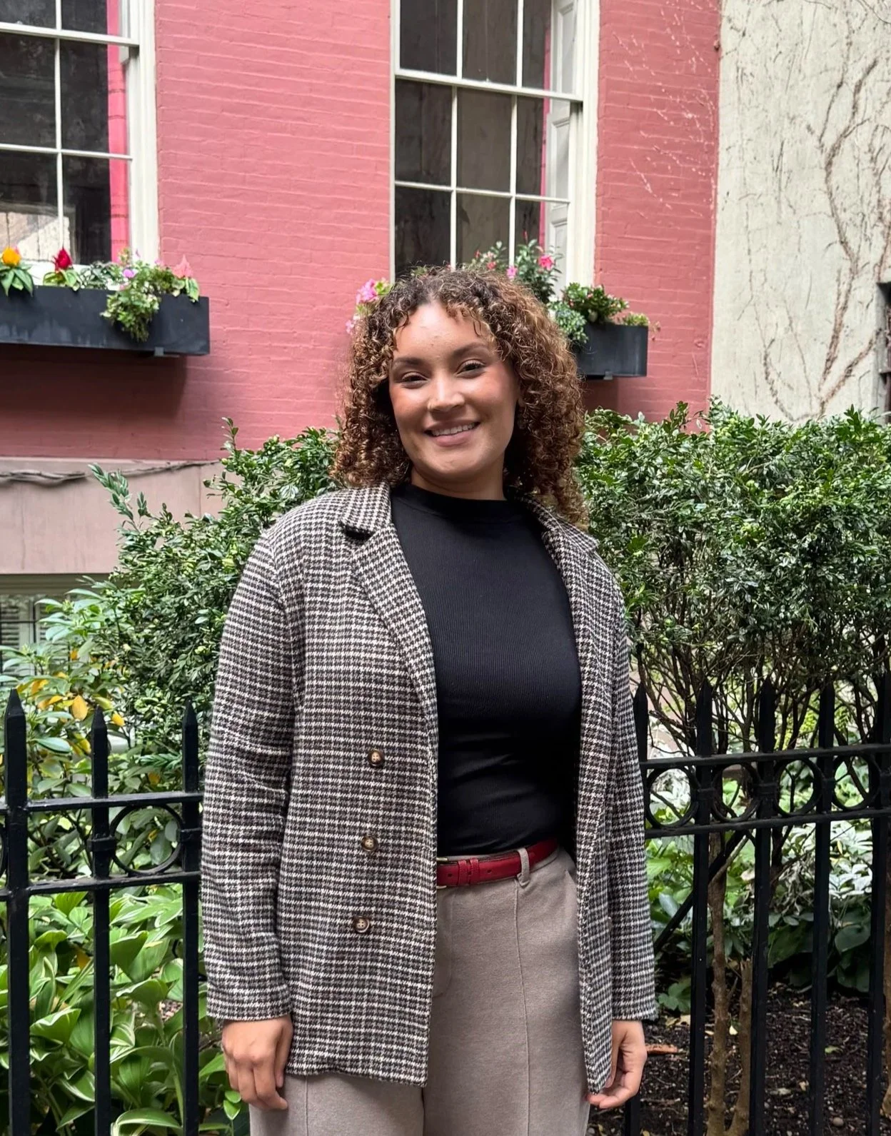 A woman with curly hair smiling outdoors in front of a bush, with a pink building and windows with window boxes filled with flowers in the background.