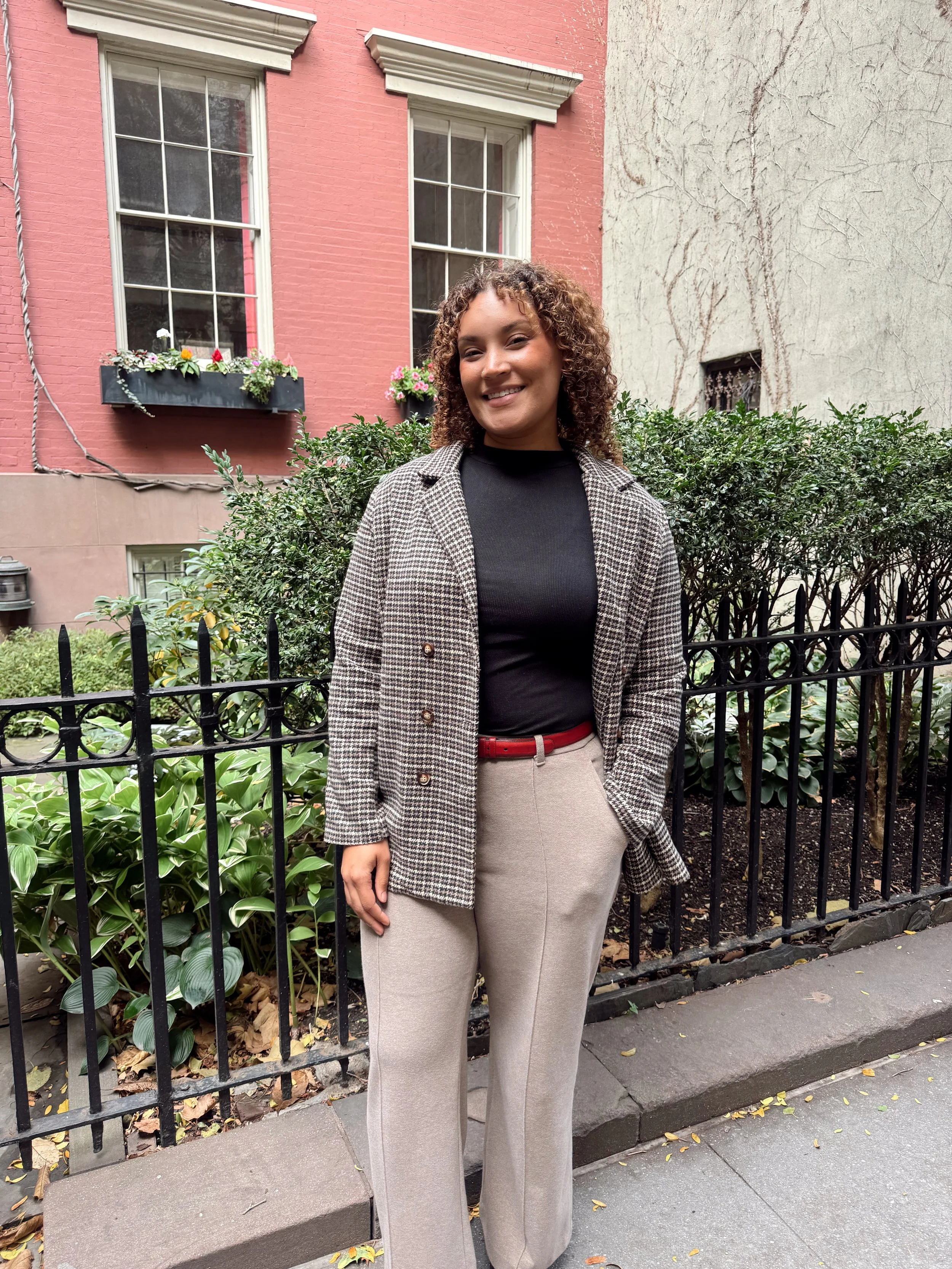 woman therapist stands on NYC street in a plaid blazer, red belt, beige pants. Her hand is in her pocket and she is smiling