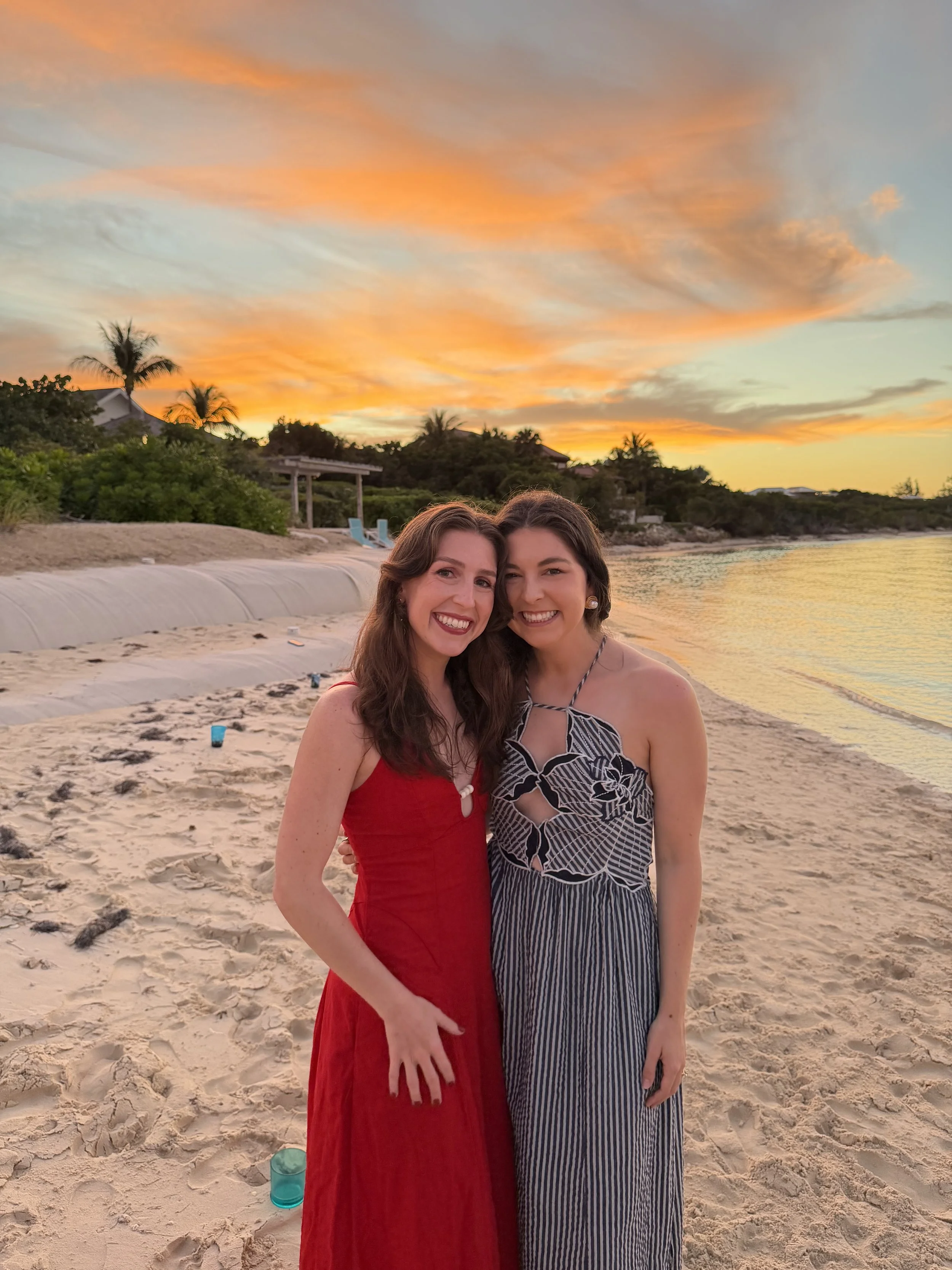 two women stand on hudson river beach with sunset behind them. One is in a red dress. the other a black and white dress.