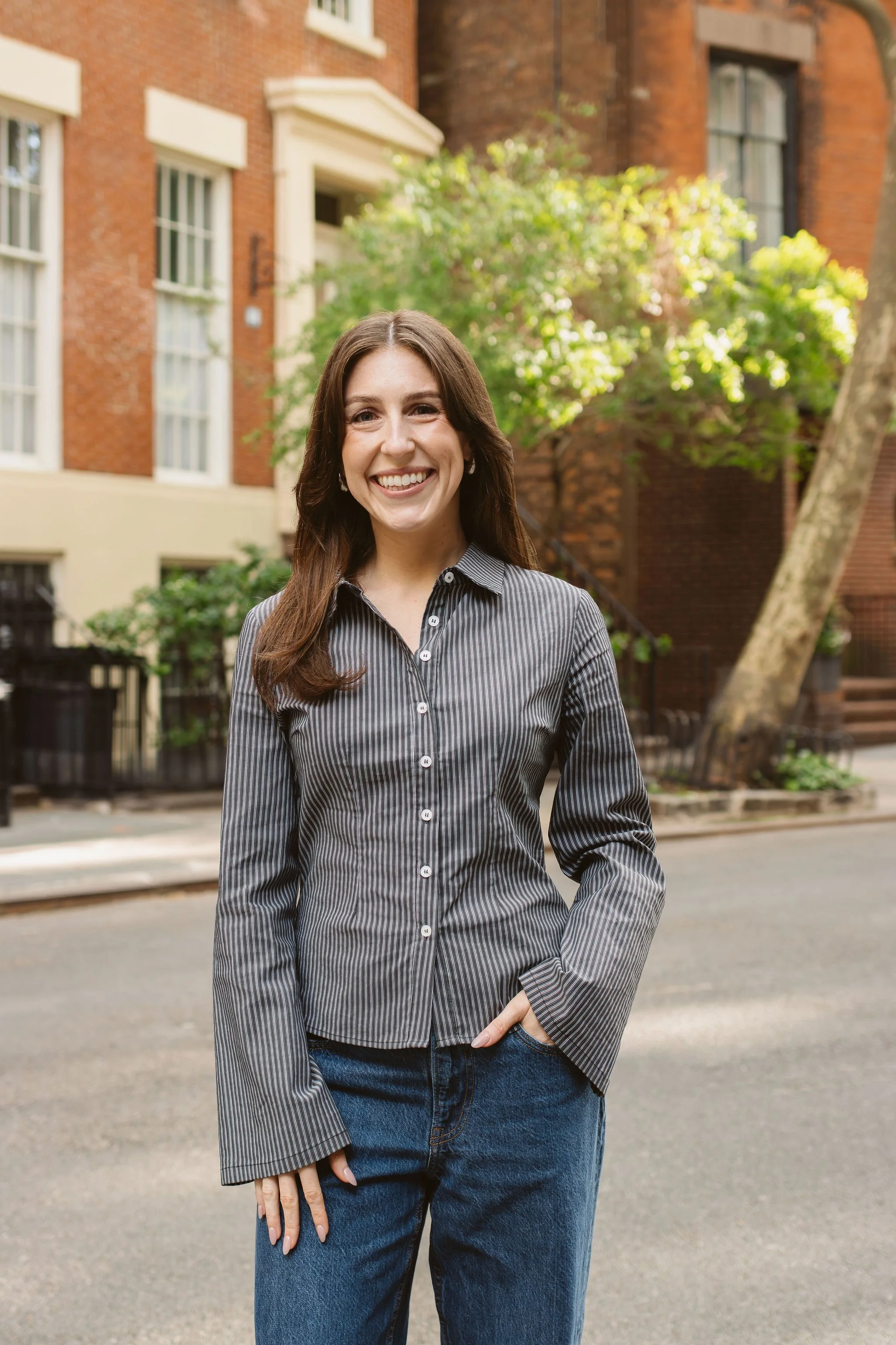 woman with straight brown hair, brown sweater, & brown plaid pants stands in front of nyc apartment building