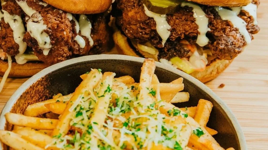 Two fried chicken sandwiches with pickles and mayonnaise on brioche buns placed on a wooden table, a bowl of French fries topped with shredded cheese and herbs, and two small cups of caramel sea salt and strawberry rhubarb dairy-free ice cream.