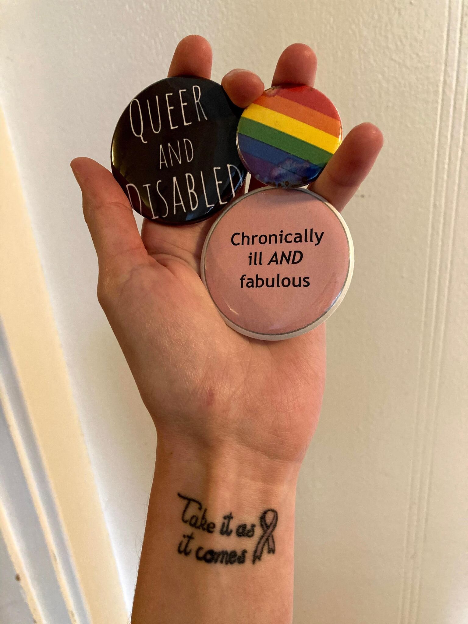 Image of Lizzie holding pins that say; “Queer and Disabled,” “Chronically Ill and Fabulous,” and the pride flag