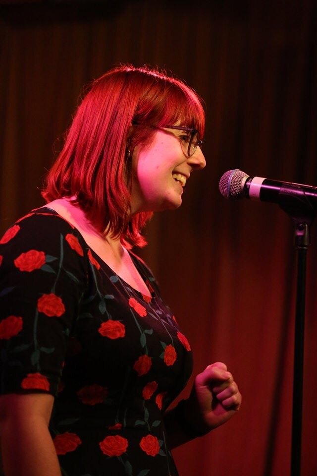 An image of the blog author in a black dress with red rose pattern speaking at a microphone.