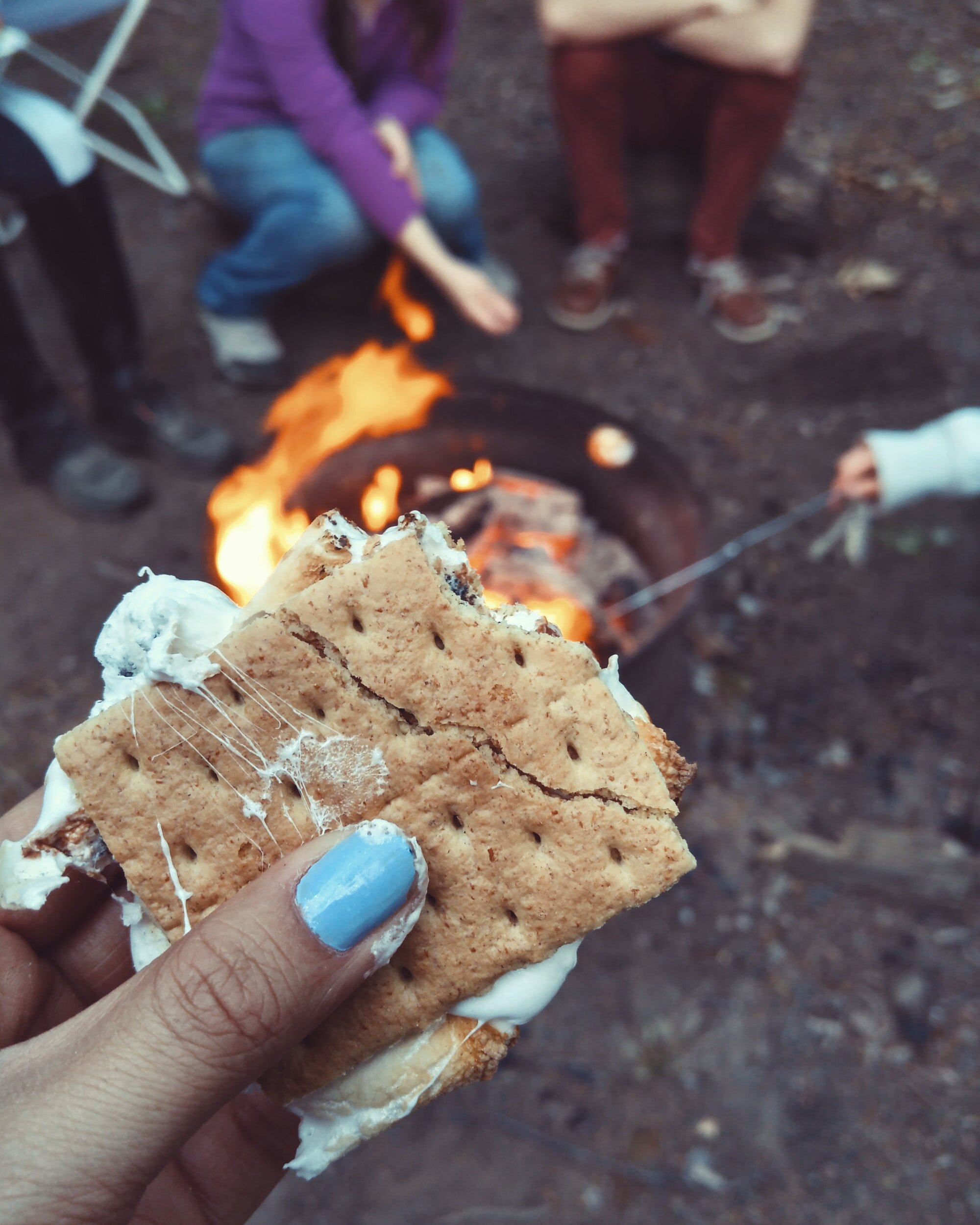 Photo of a person’s hand with blue nail polish holding a cook s’more in front of a fire by Autumn Mott Rodeheaver on Unsplash