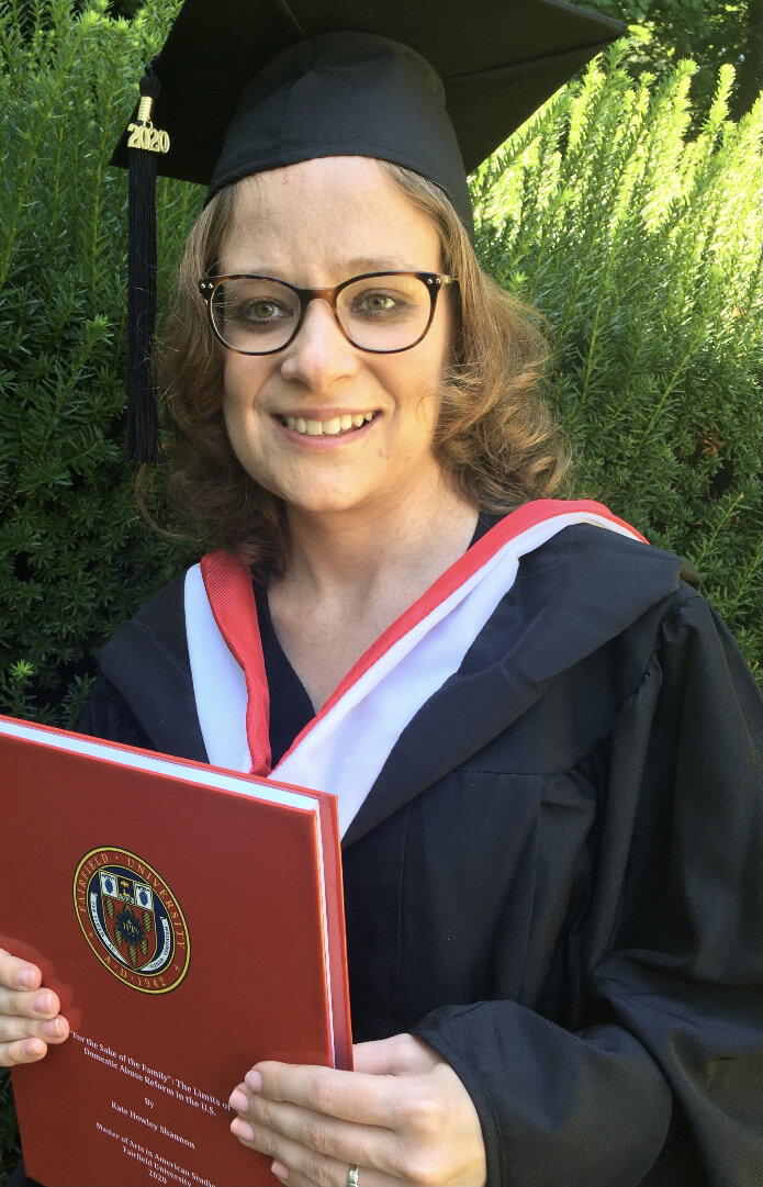 The blogger holds her MA thesis. She is standing in front of a green bush.