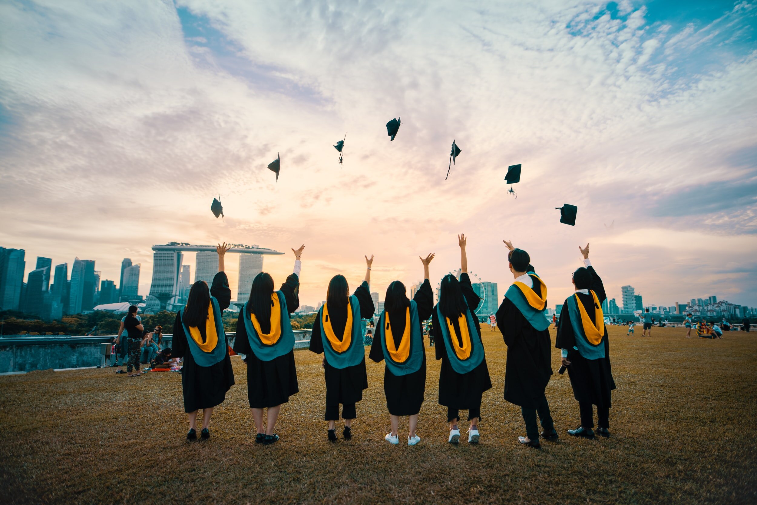 Photo by Pang Yuhao on Unsplash A group of graduates wear black robes with blue and golden hoods. They throw their caps into a cloudy sky.